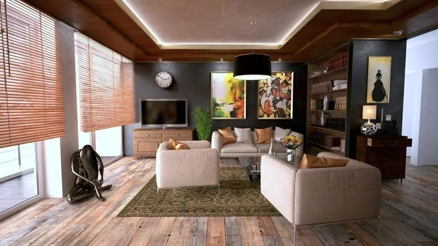 The image shows a cozy living room with wooden floors, a beige sofa, and armchairs with brown and orange pillows. The back wall features a TV, a clock, and two colorful abstract paintings that add vibrancy to the space. A bookshelf filled with books and decor items lines the right wall. A black pendant light hangs above the seating area, and large windows with wooden blinds let in natural light. The wall art, consisting of abstract paintings, enhances the room's modern aesthetic.





