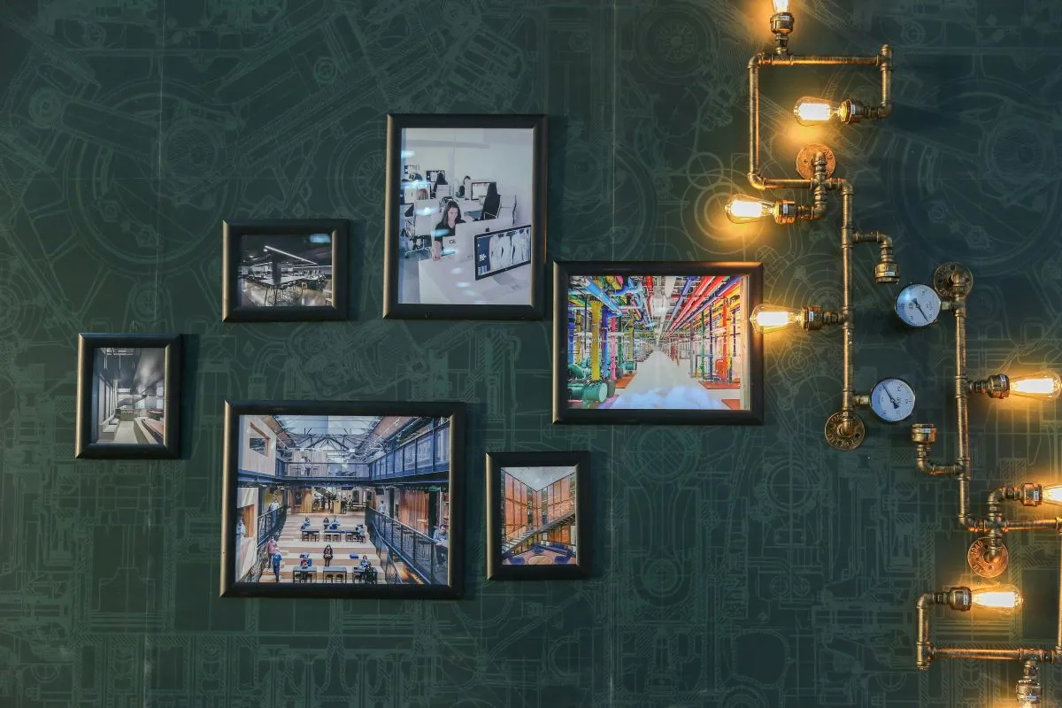 The image shows a wall with several framed photographs of industrial and office scenes, set against a dark, blueprint-patterned background. An industrial-style light fixture with brass pipes, exposed bulbs, and pressure gauges adds to the decor.