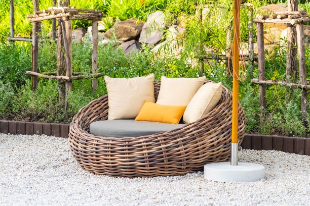 A cozy outdoor wicker lounge chair with a cushioned seat and several pillows, situated on a gravel surface, surrounded by lush greenery and wooden trellises.






