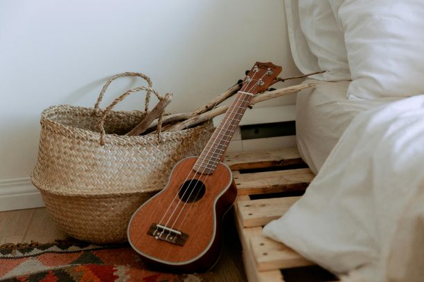 A wicker basket filled with sticks placed on a colorful rug next to a small wooden pallet bed with white bedding. A ukulele is leaning against the basket, adding a rustic and cozy touch to the scene.






