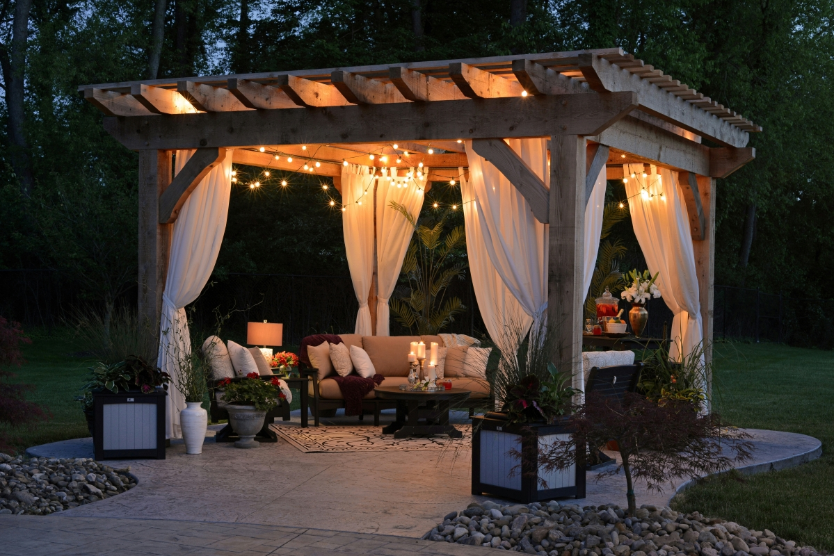 "A cozy outdoor seating area under a wooden pergola with draped white curtains, string lights, and comfortable furniture, set in a beautifully landscaped garden."





