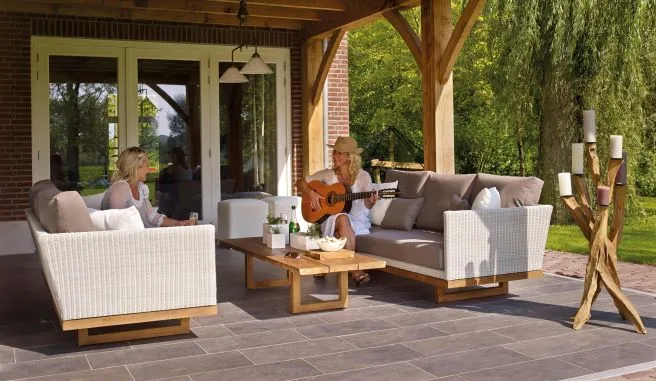 Two women enjoying a relaxed moment on a beautifully decorated garden patio. One woman is playing a guitar, while the other sips a drink. The seating area features cozy wicker sofas with plush cushions, a wooden coffee table adorned with small potted plants, and a unique candle holder made from natural wood, creating a serene and inviting atmosphere."