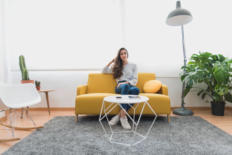 A woman is sitting on a yellow sofa in a modern, well-lit living room with a gray carpet. She has long hair, wears a striped shirt and jeans, and is looking to the side with a relaxed expression. The room features a round white coffee table, a large floor lamp, a cactus on a small table, and a large green plant. The space has a contemporary, minimalist design.