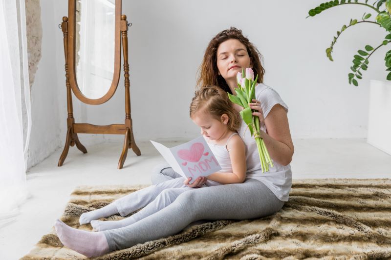 A mother and her young daughter are sitting on a soft, furry carpet in a bright, minimalist room. The mother is holding a bouquet of tulips and has her eyes closed, appearing content. The daughter is sitting in her mother's lap, focused on a card that says "Mom" with a heart drawn on it. The room features a wooden floor mirror, a large green plant, and sheer white curtains letting in natural light.