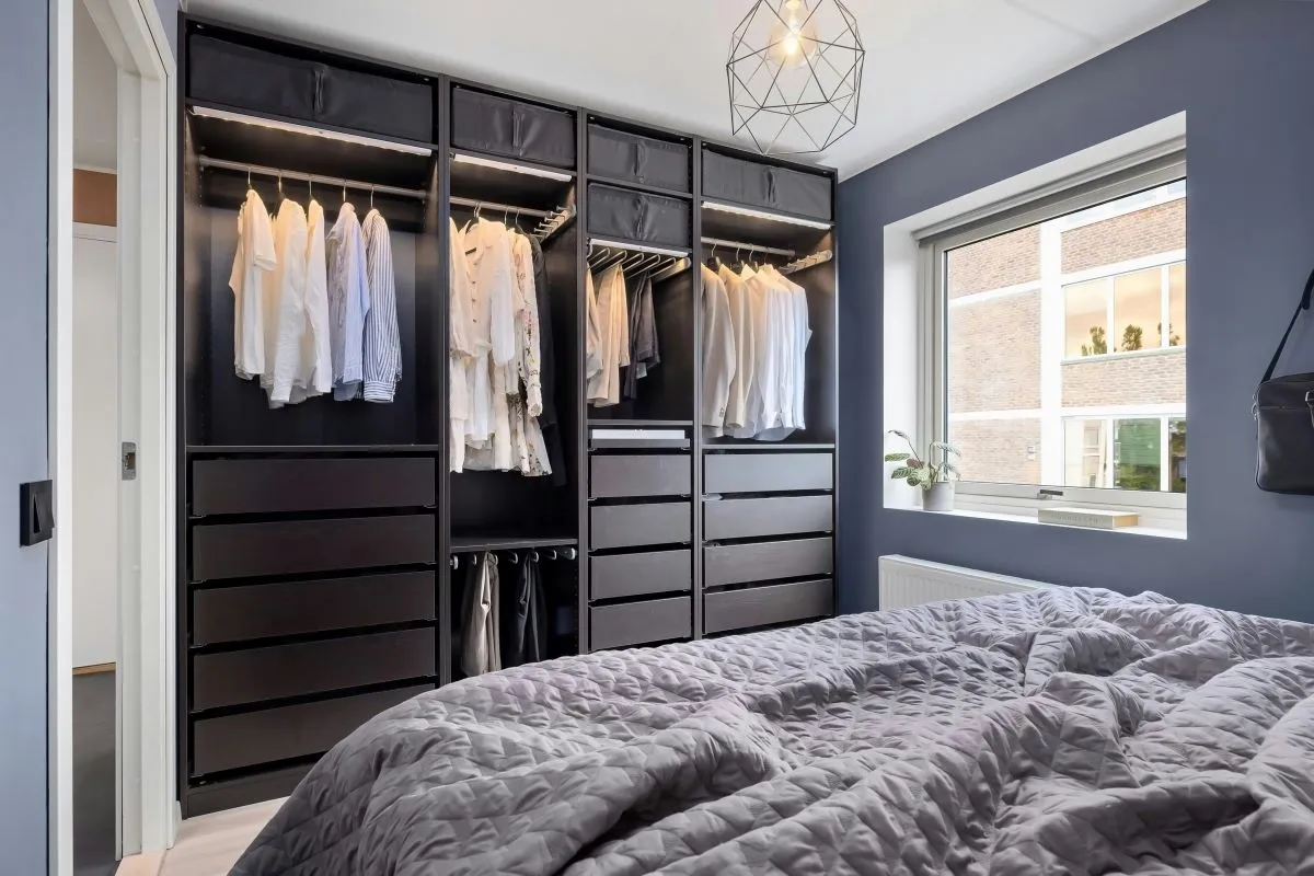 "Modern bedroom with an open black wardrobe featuring hanging clothes, multiple drawers, and storage compartments, next to a window with natural light."