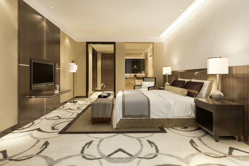 A luxurious, modern hotel room with a large bed, nightstands, stylish lamps, and a plush carpet with an abstract pattern. A flat-screen TV is mounted on a sleek wooden panel. The room opens into a spacious bathroom with modern fixtures. The décor is warm and sophisticated.