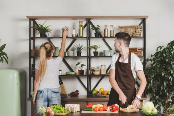 wo people in a kitchen setting, one organizing items on a stylish indoor rack and the other preparing vegetables on a counter."