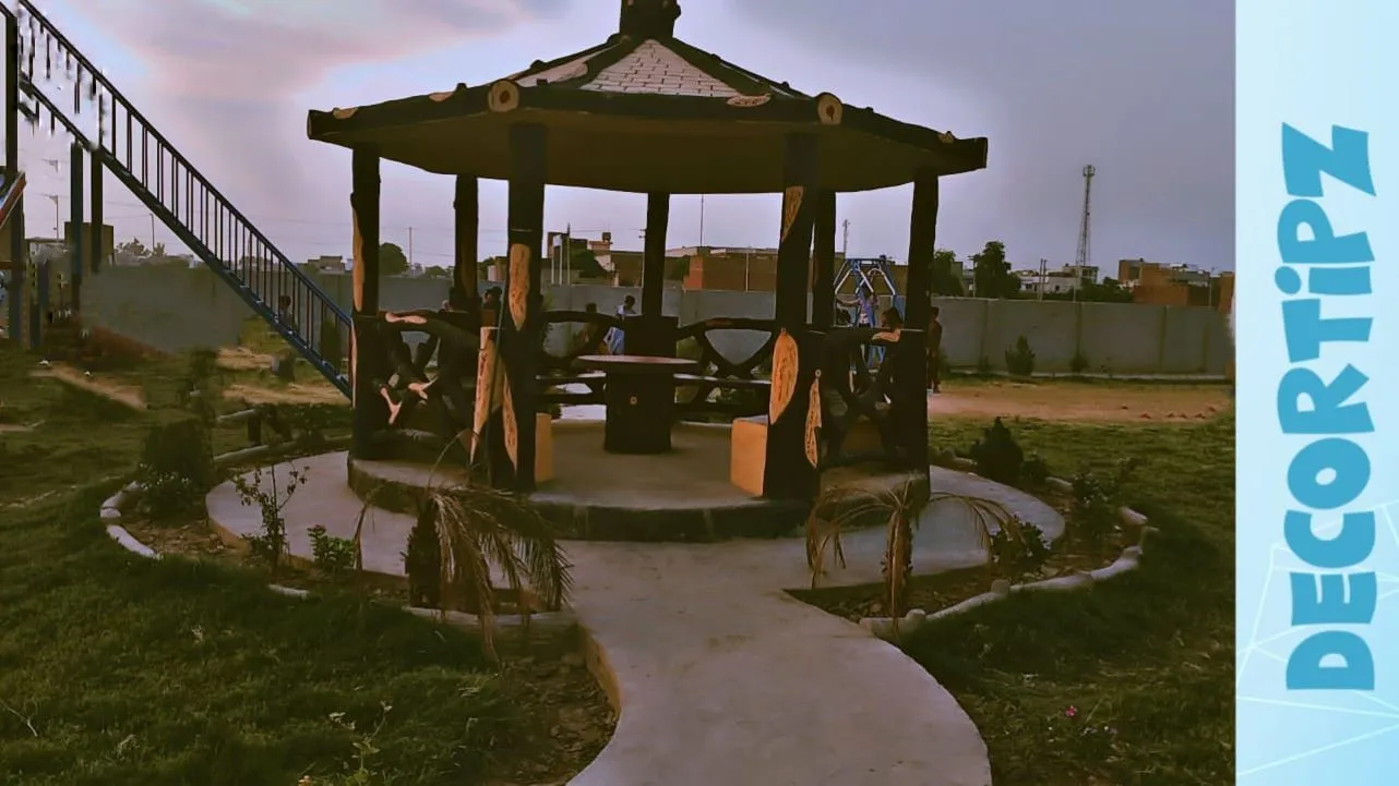 A wooden gazebo with benches and a central table, set in a home garden with surrounding greenery and a playground in the background.