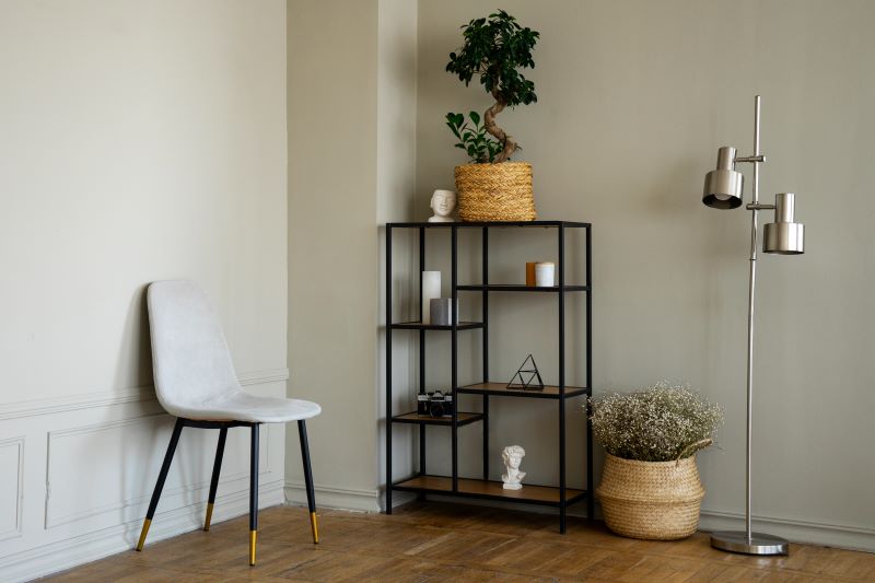 A minimalist room with a gray chair, black metal rack with decorative items, silver floor lamp, and woven baskets with plants against light gray walls and wooden flooring.