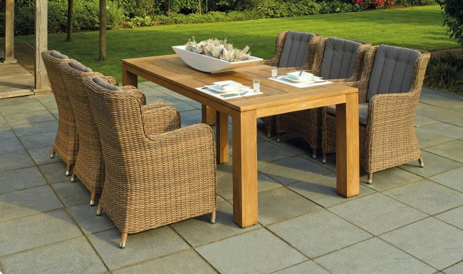 A wooden outdoor dining table with wicker chairs and cushioned seats on a tiled patio, set up for dining with plates and a decorative centerpiece, surrounded by a lush green garden.