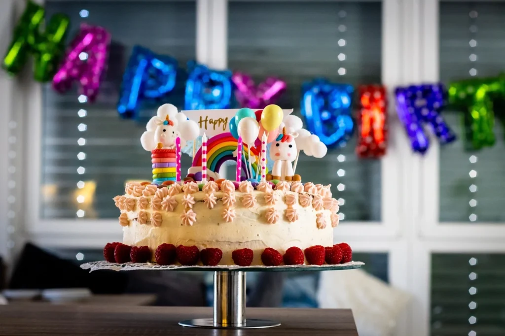 A colorful birthday cake decorated with strawberries, rainbow toppers, and unicorn figures, in a birthday party set against a festive "Happy Birthday" banner.