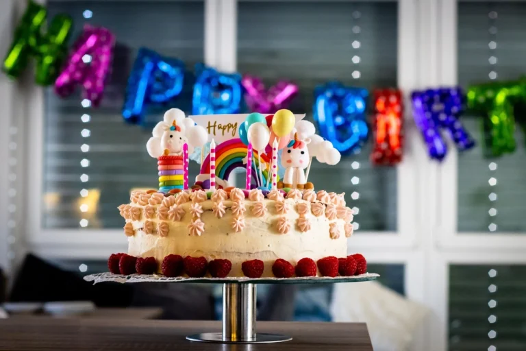 A colorful birthday cake decorated with strawberries, rainbow toppers, and unicorn figures, in a birthday party set against a festive "Happy Birthday" banner.