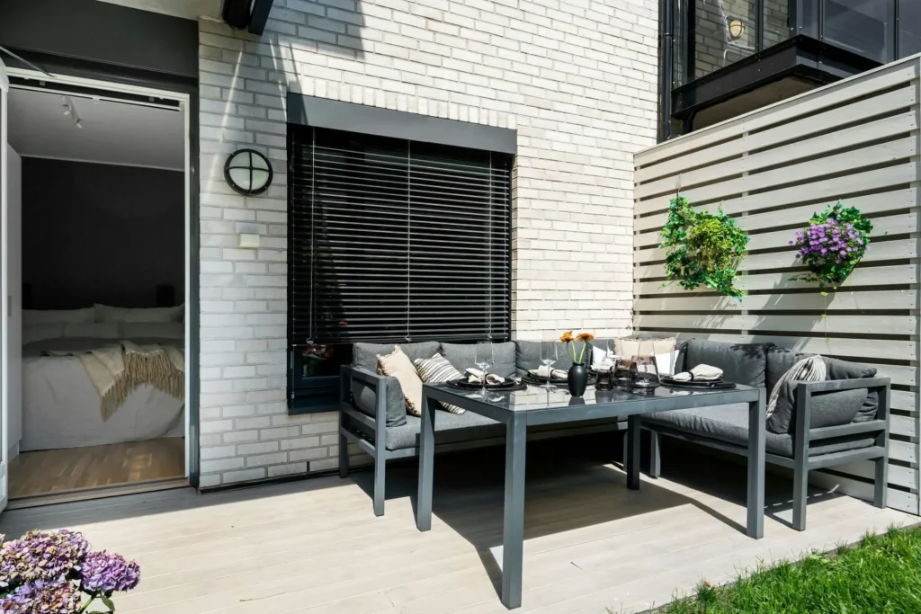 A modern outdoor patio with a corner sofa, glass-top dining table, hanging plants, and an open door leading to a bedroom, all set against light brick walls with a minimalist design.