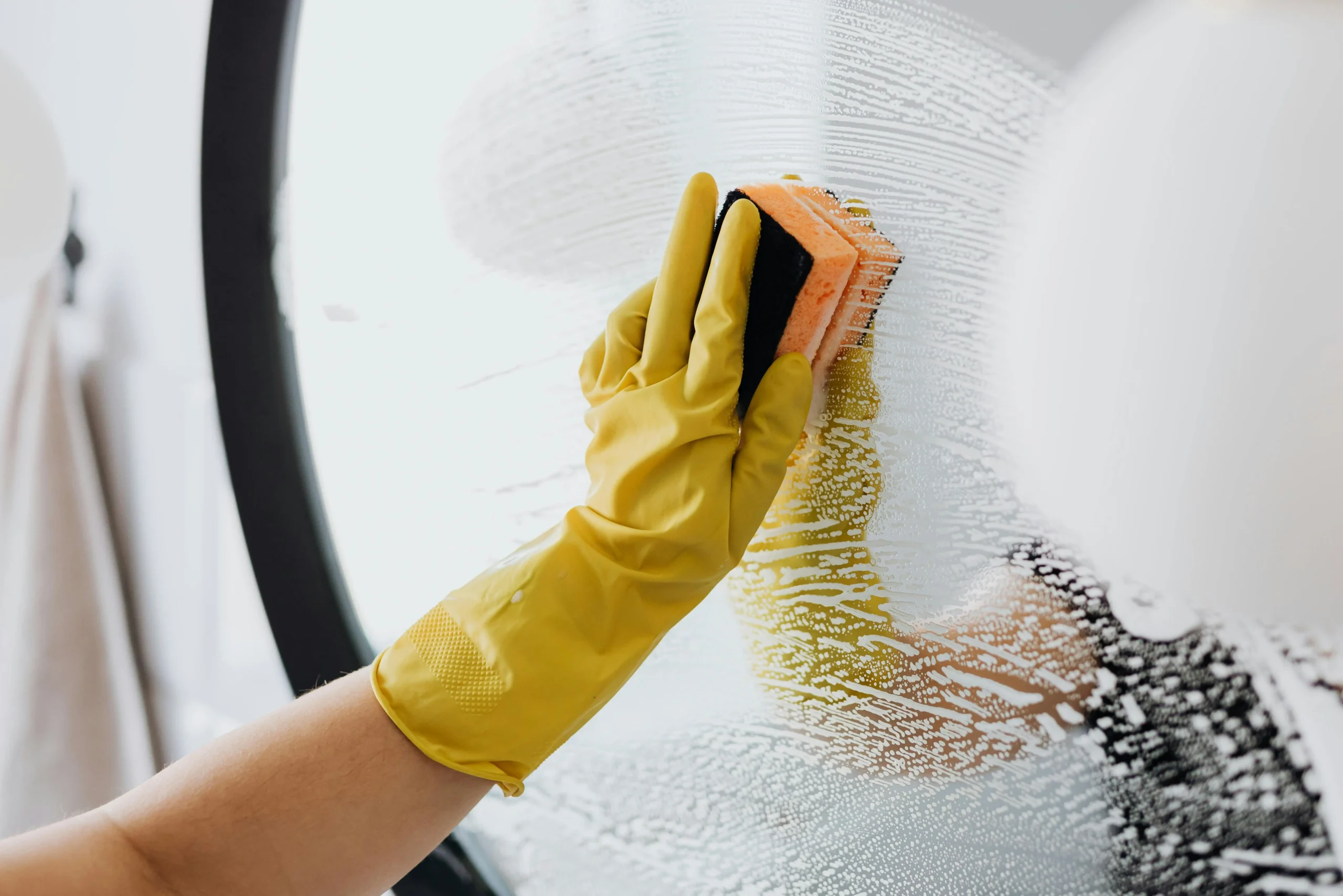 Hand wearing a yellow glove scrubbing a soapy mirror, demonstrating essential spring cleaning tips for a sparkling home.