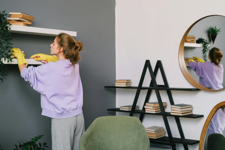 Woman dusting a bookshelf in a modern living room, demonstrating spring cleaning tips for a tidy home.