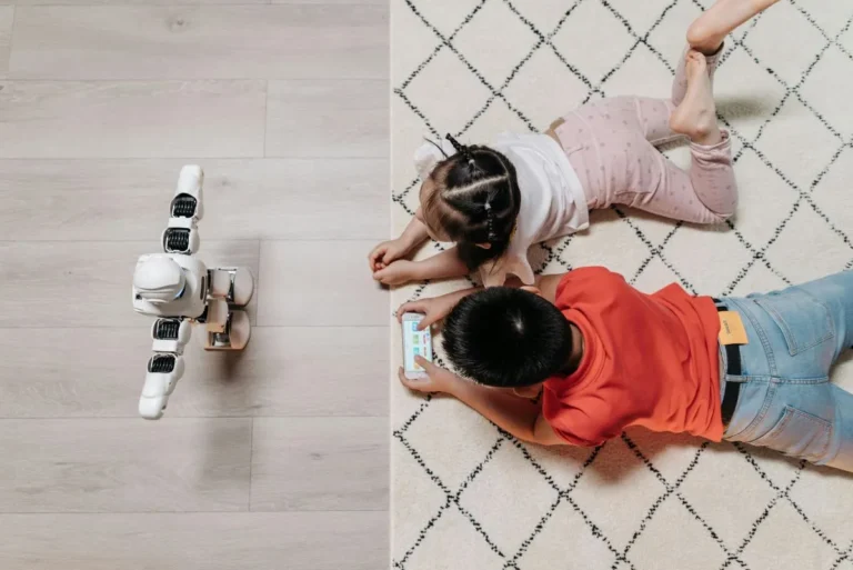 A small AI-powered robot interacts near two children lying on the floor, one watching a smartphone, highlighting modern smart home technology.