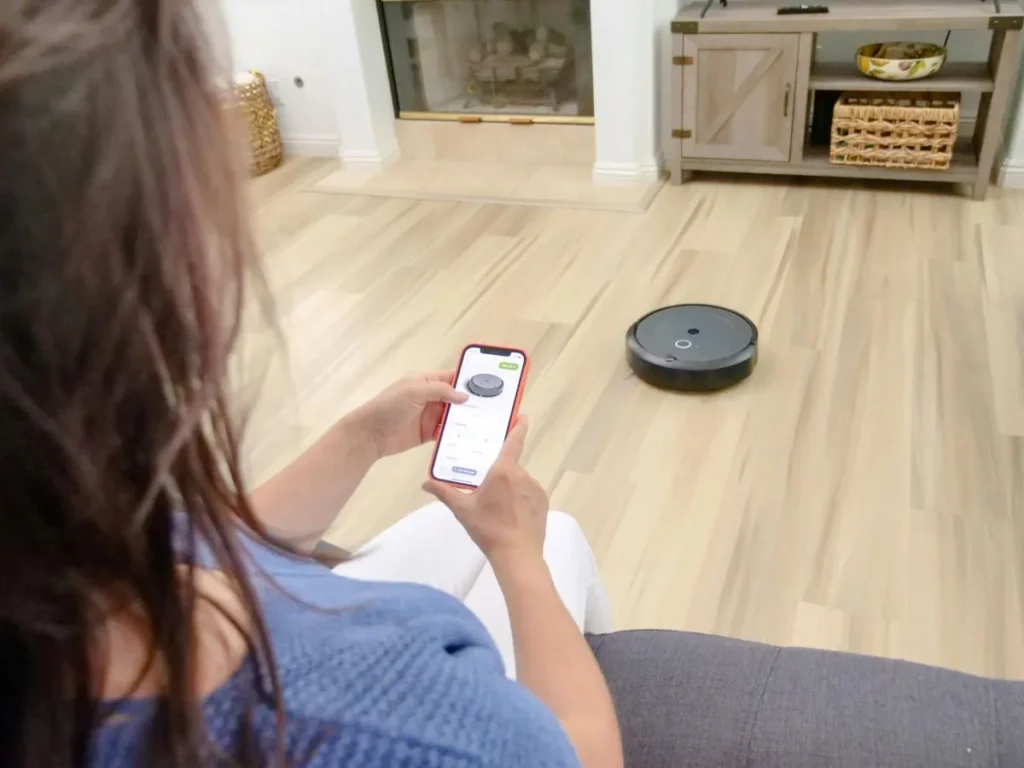 A woman is sitting in her living room, controlling a robot vacuum cleaner using an app on her smartphone. The robot vacuum is moving across the wooden floor while she adjusts the settings on her device.