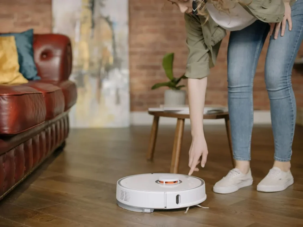 "A person in casual clothing is pressing a button on a white robotic vacuum cleaner on a wooden floor in a modern living room with a leather sofa, a small wooden table, and a potted plant in the background."