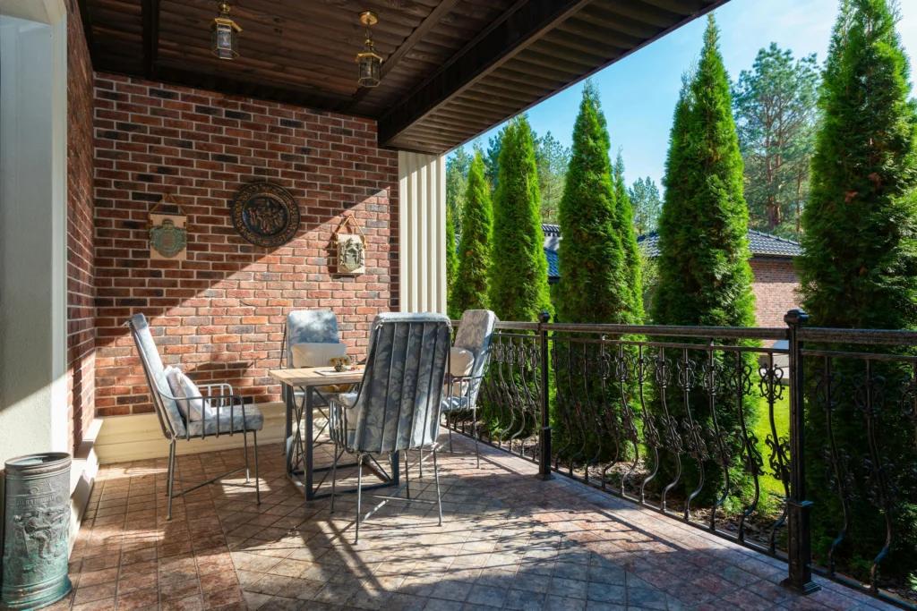Outdoor patio seating area with metal garden chairs and table on a covered balcony, featuring brick walls, decorative railing, and green trees in the background.