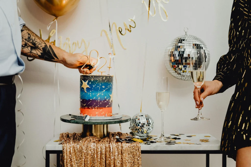 Elegant New Year home decoration showing a decorated table with a celebration cake, balloons, disco balls, and champagne glasses.