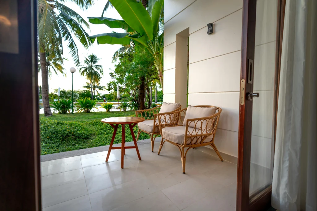 Cozy outdoor patio with wicker chairs and a small wooden table, overlooking a green garden with palm trees, ideal for relaxing outdoor seating.